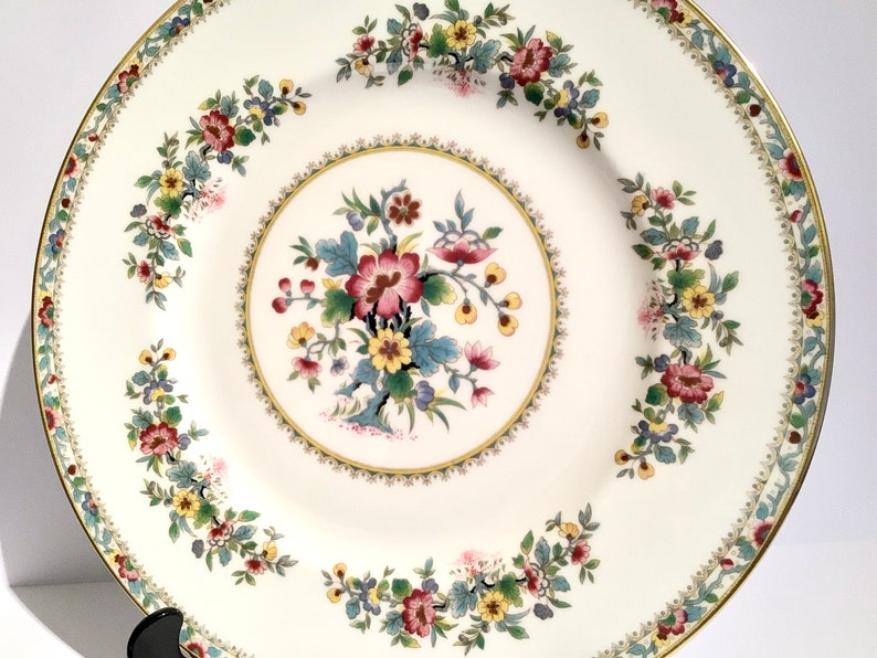 May include: A white porcelain dinner plate with a gold rim and a floral design. The center of the plate features a large floral motif with pink, yellow, and blue flowers. The rim of the plate is decorated with a smaller floral pattern.
