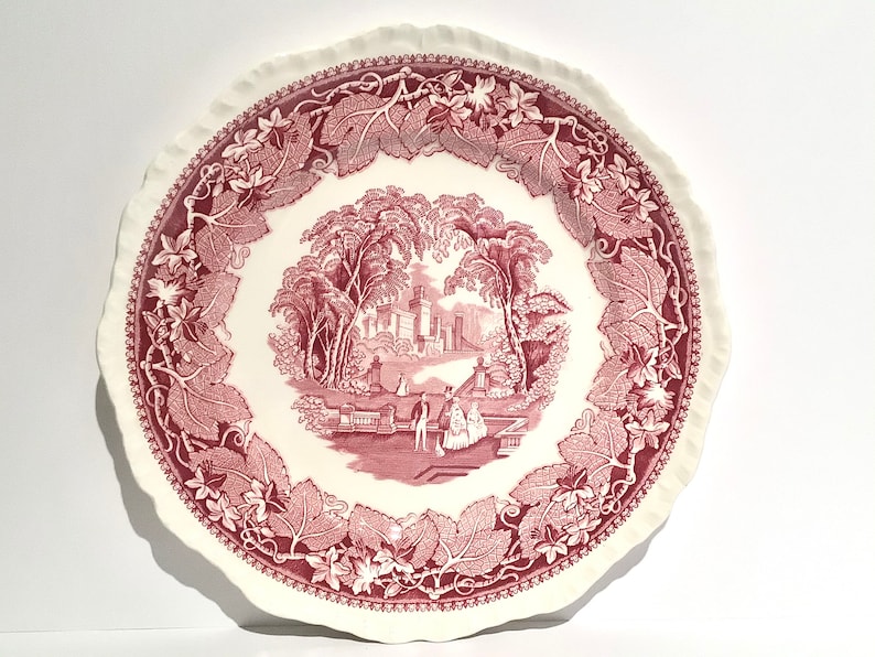 May include: A pink and white ceramic dinner plate with a scalloped edge. The plate features a pink floral design with a central image of a garden scene with a building and people.