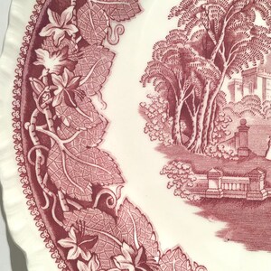 May include: A vintage ceramic plate with a pink and white floral design. The plate features a detailed border of leaves and vines, and a central image of a castle with trees and a couple walking in the foreground.