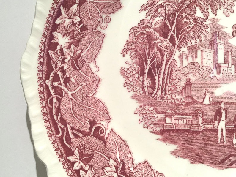 May include: A vintage ceramic plate with a pink and white floral design. The plate features a detailed border of leaves and vines, and a central image of a castle with trees and a couple walking in the foreground.