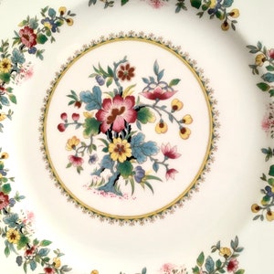 May include: A white ceramic dinner plate with a floral design. The plate has a gold rim and a center design of a large pink flower with yellow and blue accents. The border of the plate features a repeating pattern of smaller flowers in shades of pink, yellow, and green.