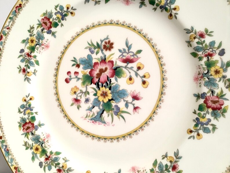 May include: A white ceramic dinner plate with a floral design. The plate has a gold rim and a center design of a large pink flower with yellow and blue accents. The border of the plate features a repeating pattern of smaller flowers in shades of pink, yellow, and green.
