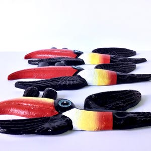 May include: A collection of wooden toucan ornaments. Each toucan features a vibrant red beak, a black body, and accents of white, yellow, and red. The toucans are displayed in a row, highlighting their colourful details. Ideal for home decoration.