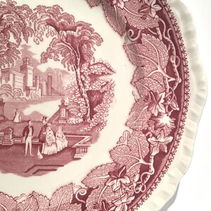 May include: A vintage pink and white ceramic plate with a detailed floral border and a central image of a castle and figures in a garden.