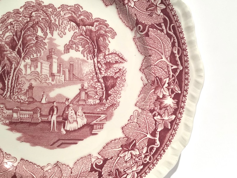May include: A vintage pink and white ceramic plate with a detailed floral border and a central image of a castle and figures in a garden.