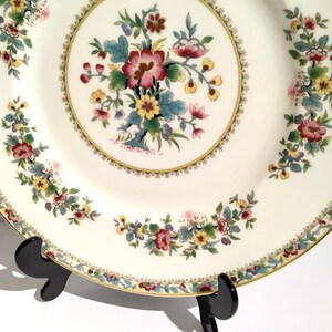 May include: A white porcelain dinner plate with a gold rim and a floral design in shades of pink, yellow, green, and blue. The plate is on a black stand.
