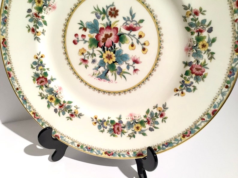 May include: A white porcelain dinner plate with a gold rim and a floral design in shades of pink, yellow, green, and blue. The plate is on a black stand.