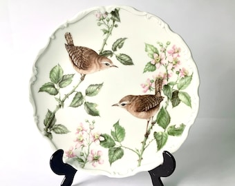 May include: A decorative white plate with a scalloped edge, featuring two brown birds perched on branches with green leaves and pink flowers. The plate is supported by a black stand.