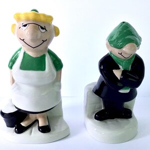 May include: Two ceramic figurines depicting a pair of characters. The left figure has yellow hair, a green top, and a white apron. The right figure wears a dark blue coat, a green scarf, and a green cap. Both figures stand on white bases.