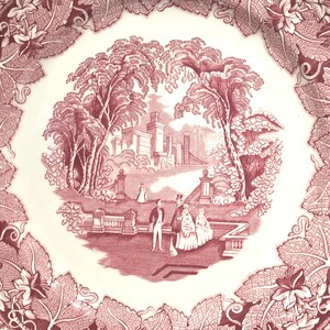 May include: A vintage pink and white ceramic plate with a detailed design of a manor house surrounded by trees and a couple walking in a garden. The plate has a scalloped edge and a vine border.