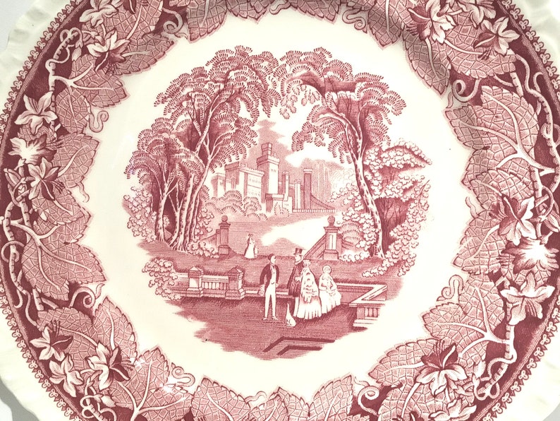 May include: A vintage pink and white ceramic plate with a detailed design of a manor house surrounded by trees and a couple walking in a garden. The plate has a scalloped edge and a vine border.