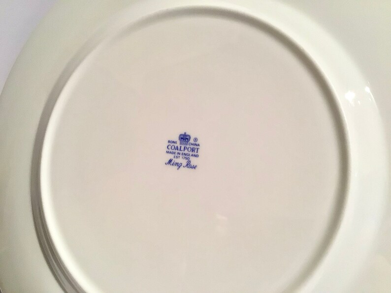 May include: The bottom of a white ceramic plate with a blue and white logo that reads "Bone China Coalport Made in England Est 1750 Ming Rose"