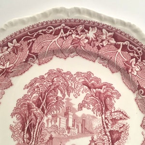 May include: A vintage ceramic plate with a pink and white floral design. The plate features a detailed illustration of a castle surrounded by trees and vines.