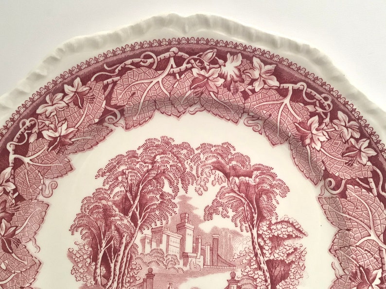 May include: A vintage ceramic plate with a pink and white floral design. The plate features a detailed illustration of a castle surrounded by trees and vines.