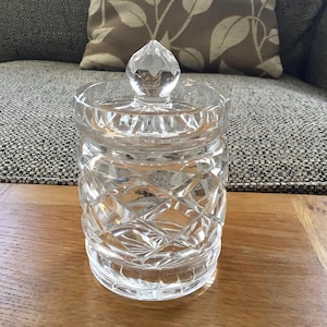 Könnte beinhalten: Ein klarer, geschliffener Glasbehälter mit Deckel, der auf einer Holzoberfläche steht. Der Behälter hat ein Rautenmuster und einen facettierten Knauf oben. Der Hintergrund umfasst ein graues Sofa und ein gemustertes Kissen.