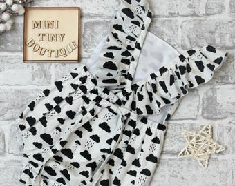 May include: A white baby onesie with black clouds and raindrops. The onesie has ruffled straps and a tie at the back. The onesie is on a white brick background. The text "MINI TINY BOUTIQUE" is on a wooden sign in the top left corner of the image.
