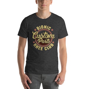 Pode incluir: Uma camiseta cinza escura com um logotipo de estilo vintage que diz "Bionic Custom Parts Knee Club". O logotipo está em um círculo com estrelas ao redor e as palavras estão em uma fonte amarela e marrom.