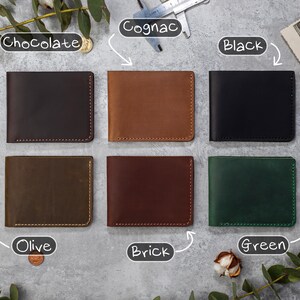 May include: Six leather wallets in different colors: chocolate, cognac, black, olive, brick, and green. Each wallet has a stitched edge.