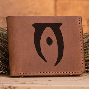 May include: A brown leather wallet with a stitched border. The wallet features a dark brown graphic design on the front. The design is a stylized symbol with a central oval shape.