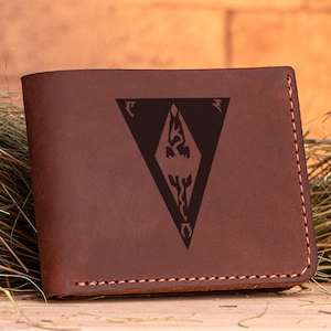 May include: A brown leather wallet with a stitched border. The wallet features a dark brown, stylized emblem in the center. The wallet is rectangular and appears to be made of high-quality leather.