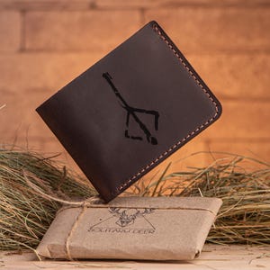 May include: A brown leather wallet with a black stitched edge and a black graphic of a stylized tree with branches. The wallet is floating above a pile of dried grass.