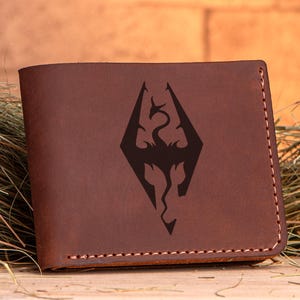 May include: A brown leather wallet with a stitched border. The wallet features a black graphic of a dragon within a diamond shape. The wallet is rectangular and appears to be new.