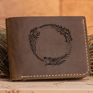 May include: Brown leather wallet with a dragon emblem. The rectangular wallet has a stitched edge. The dragon design is a circular emblem with two dragons facing each other. The wallet is on a wooden surface.