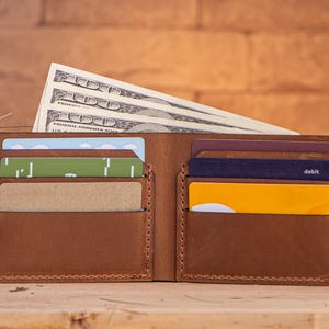 May include: A brown leather wallet with multiple card slots and a cash compartment. The wallet is open and contains cash and credit cards. The wallet is sitting on a wooden surface.