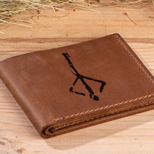 May include: A brown leather wallet with a black symbol engraved on the front. The symbol is a stylized representation of a tree with branches.