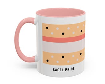 May include: A white ceramic mug with a pink interior and handle, featuring a graphic of a bagel with cream cheese and poppy seeds. The design includes horizontal bands of tan, pink, and white, with the black text BAGEL PRIDE printed below.
