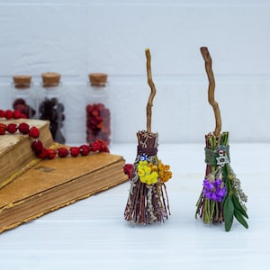 May include: Two decorative miniature brooms with wooden handles and bundles of dried flowers and twigs. One broom features yellow flowers and an evil eye charm, the other has purple flowers and a Star of David charm. The brooms are on a white surface.