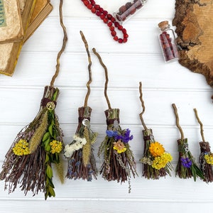 Op de afbeelding: Een set van zes miniatuurbezems gemaakt van twijgen en versierd met gedroogde bloemen en kruiden. De bezems zijn vastgebonden met touw en hebben elk een klein sieraad.