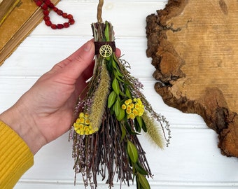 May include: A decorative miniature broom made of twigs, dried flowers, and greenery. The broom features a brown handle and is adorned with yellow, green, and purple floral accents. A gold-coloured charm with a tree of life design is attached.