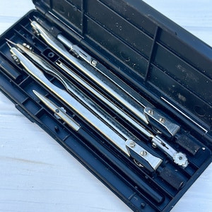 May include: A black plastic case containing a set of four silver compasses. The compasses are closed and stored inside the case.