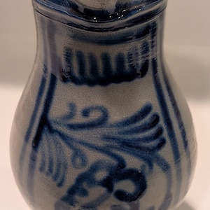 May include: A ceramic pitcher with a light gray base and a cream-colored rim. The pitcher features blue floral and geometric designs. The rim is accented with a blue band. The pitcher has a spout for pouring.