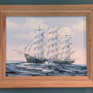 May include: A framed oil painting of a large sailing ship with white sails, navigating a blue ocean under a cloudy sky.