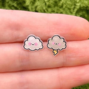 May include: A pair of cloud-shaped earrings. One earring features a smiling cloud with pink cheeks, the other has a lightning bolt. The earrings have a black outline and are set against a green background.