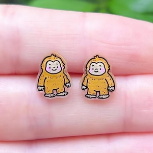 May include: A pair of Bigfoot stud earrings. Each earring depicts a cartoon yellow Bigfoot with a smiling face and pink cheeks. The earrings are small and appear to be made of a clear material.