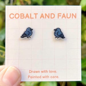 Pode incluir: Um par de brincos em forma de pássaro azul cobalto em um cartão branco com o texto "COBALT AND FAUN". O cartão também diz "Drawn with love. Painted with care."