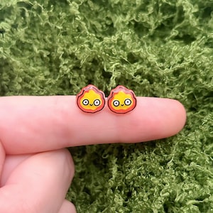 Può includere: Un paio di piccoli orecchini in stile cartone animato con un design a fiamma giallo, arancione e nero. Gli orecchini rotondi e lucidi sono mostrati su un dito contro uno sfondo verde strutturato.