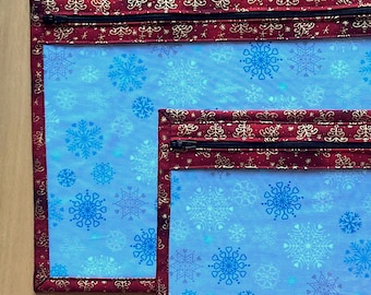 May include: Two rectangular zippered pouches. The pouches have a light blue background with a snowflake pattern. The borders are a deep red with a gold design. The zippers are black.