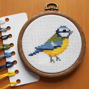 May include: A bluebird cross-stitch embroidery in a wooden hoop with a white background. The bird has a blue head, yellow breast, and green back. The embroidery is surrounded by a white card with holes for floss.