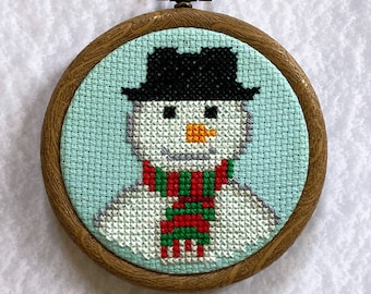 May include: A framed cross-stitch of a snowman wearing a black hat and a red and green scarf. The background is a light blue color.