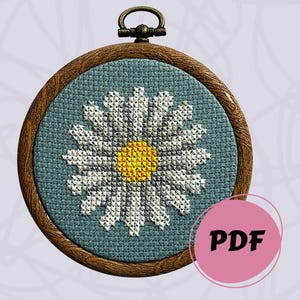 Può includere: Un telaio da ricamo circolare in legno contenente un motivo a punto croce di una margherita bianca con un centro giallo brillante su uno sfondo di tessuto blu polvere. Un piccolo anello di metallo per appendere è fissato in alto. Un'icona rosa circolare mostra PDF.