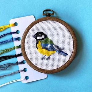 May include: A close-up of a cross-stitch embroidery of a blue tit bird on a white background. The bird is surrounded by a brown wooden frame.