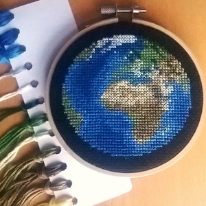 K&ouml;nnte beinhalten: Nahaufnahme einer handgestickten Kreuzstich-Stickerei der Erde in einem runden Holzstickrahmen. Die Stickerei ist in Blaut&ouml;nen, Gr&uuml;n, Braun und Wei&szlig; gehalten.