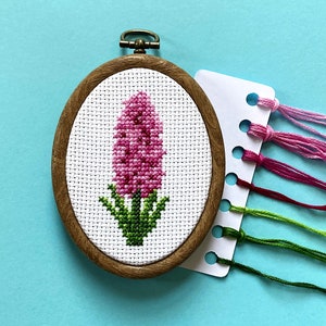 May include: Oval wooden embroidery hoop with a cross-stitch of a pink flower. Green leaves and stem are at the base. A card with pink and green embroidery floss is next to the hoop. The floss is in various shades.