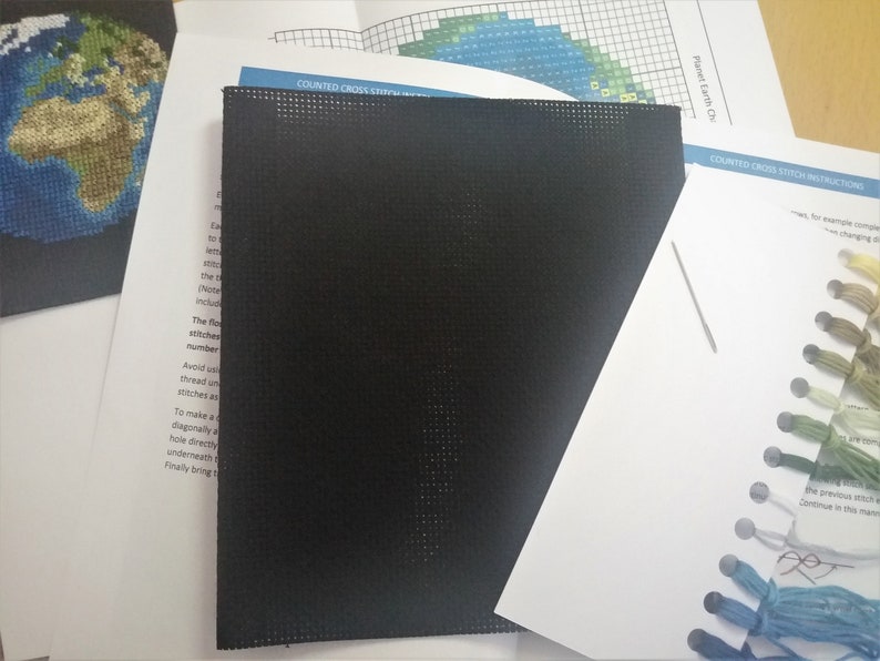 May include: A black cross stitch Aida cloth with a needle and a variety of colored embroidery floss. The instructions for the counted cross stitch project are visible in the background. The project is titled "Planet Earth Cross Stitch".