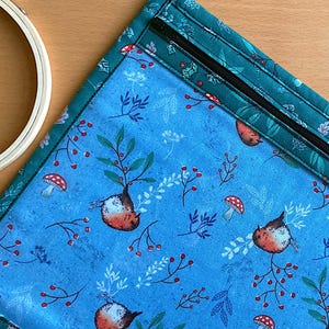 Könnte beinhalten: Eine blaue Reißverschlusstasche mit grünem Rand und einem sich wiederholenden Muster aus roten Beeren, grünen Blättern und kleinen Vögeln. Die Tasche ist aus Stoff und hat einen Reißverschluss.