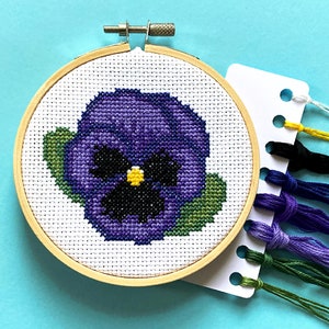 以下が含まれることがあります： 木製の刺繍枠に、紫と緑のパンジーの花のクロスステッチ刺繍が施されています。刺繍は、さまざまな色の糸のサンプルがついた白いカードで囲まれています。